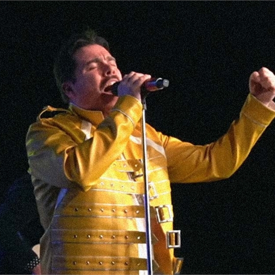 A photo of the singer of Queen Will Rock You, a tribute band, wearing a yellow jacket and singing into a microphone
