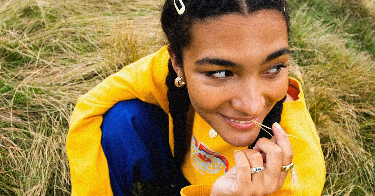 Olivia Dean sat on the grass smiling, wearing blue jeans and a yellow sweater