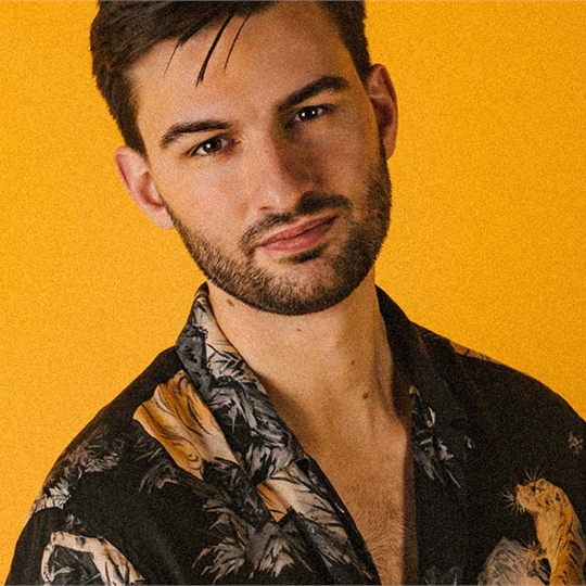 Tom One stood in front of a yellow backdrop wearing a floral shirt