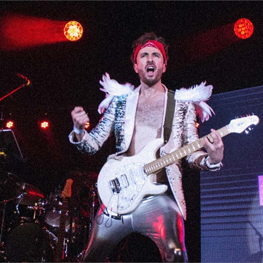 A person on a stage wearing a white, glittery, feathery, shiny suit and holding a white guitar